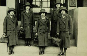 Five women pose on steps, holding black bags and wearing matching coats and hats
